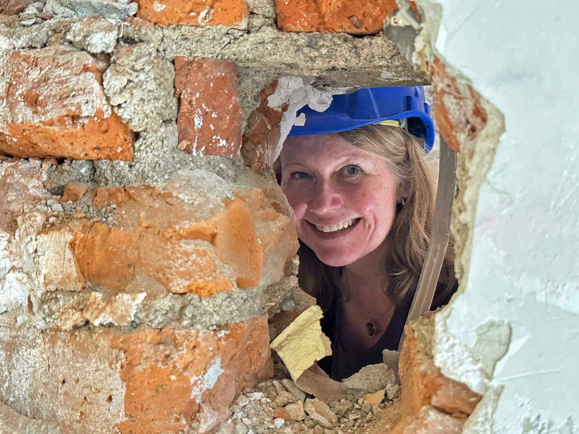 Die Architektin mit Helm auf dem Kopf lächelt durch ein Loch in der Wand in die Kamera