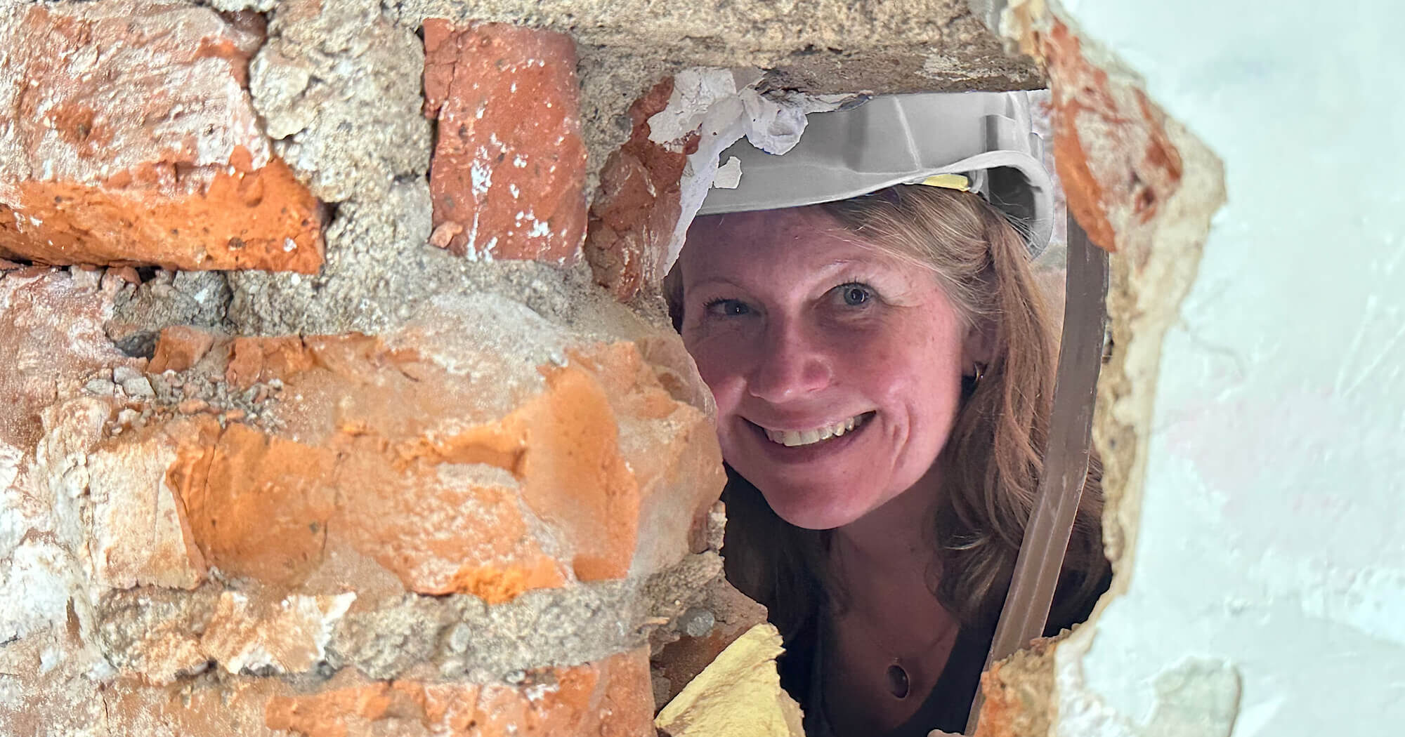 Die Architektin mit Helm auf dem Kopf lächelt durch ein Loch in der Wand in die Kamera